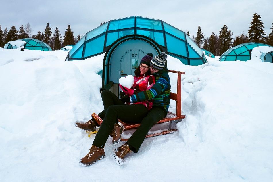 Aurelia Teslaru and Dan Moldovan sitting on a wooden sled in front of a glass igloo during winter at Kakslauttanen Arctic Resort Lapland